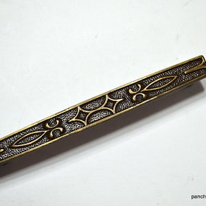 May include: A rectangular, antique brass drawer pull with an ornate design. The pull features a textured, dark background with raised, gold-colored floral and starburst patterns. The overall design is symmetrical and detailed.