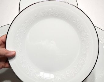 May include: A round white ceramic dinner plate featuring a subtle embossed floral pattern along the rim and a thin silver metallic band around the outer edge. The plate has a smooth, glossy finish and a clean, minimalist design for table settings.