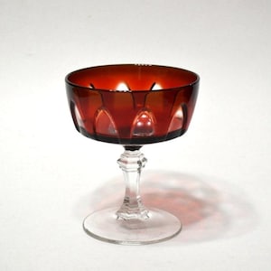 May include: A ruby red vintage champagne coupe glass. The bowl has a cut design, and the stem is clear with a faceted design. The base is a clear, round disc. The glass is on a white background.