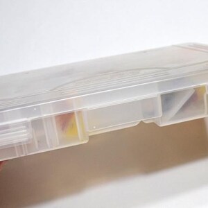 May include: A clear plastic storage box with a hinged lid and multiple compartments. The box is held in a hand and appears to contain small items. The box is rectangular and has a translucent appearance. The website address "panchosporch.etsy.com" is visible.