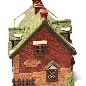 May include: A ceramic miniature house with a red brick facade, green roof, and three chimneys. The house has small windows and a decorative fence. Snow-like accents are visible on the roof and around the base. The house is a decorative item.