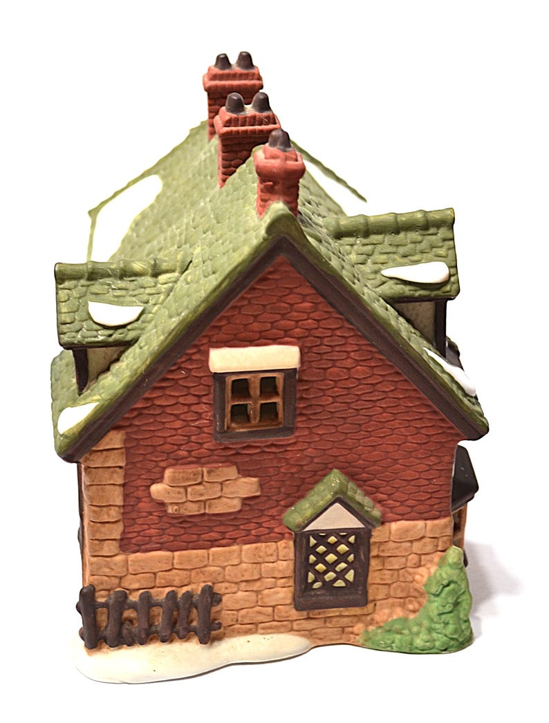 May include: A ceramic miniature house with a red brick facade, green roof, and three chimneys. The house has small windows and a decorative fence. Snow-like accents are visible on the roof and around the base. The house is a decorative item.