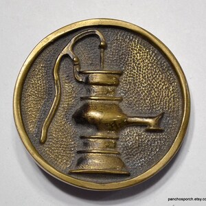 K&ouml;nnte beinhalten: Runder, antiker Messingfarbener Metallknopf mit erhabenem Reliefdesign einer Wasserpumpe. Die Pumpe ist auf einem strukturierten Hintergrund in einem kreisf&ouml;rmigen Rahmen zentriert. Der Knopf hat eine Vintage-&Auml;sthetik.