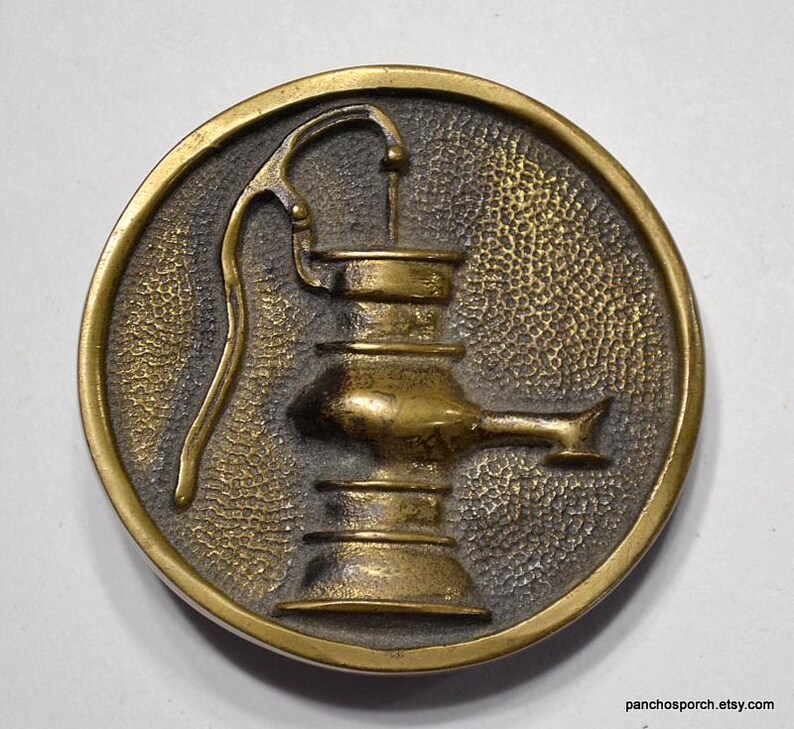 K&ouml;nnte beinhalten: Runder, antiker Messingfarbener Metallknopf mit erhabenem Reliefdesign einer Wasserpumpe. Die Pumpe ist auf einem strukturierten Hintergrund in einem kreisf&ouml;rmigen Rahmen zentriert. Der Knopf hat eine Vintage-&Auml;sthetik.