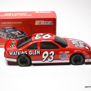 May include: A red die-cast NASCAR race car model with the number 93 on the side and top. The car has black tires and sponsor decals. The box behind the car is red and features a picture of a race car.