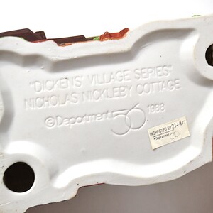 May include: The underside of a ceramic "Dickens' Village Series" Nicholas Nickleby Cottage figurine. The base is white with black holes and text that reads "Dickens' Village Series" and "Nicholas Nickleby Cottage" and "1988".