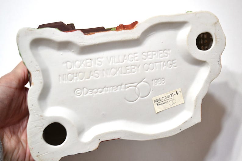 May include: The underside of a ceramic "Dickens' Village Series" Nicholas Nickleby Cottage figurine. The base is white with black holes and text that reads "Dickens' Village Series" and "Nicholas Nickleby Cottage" and "1988".