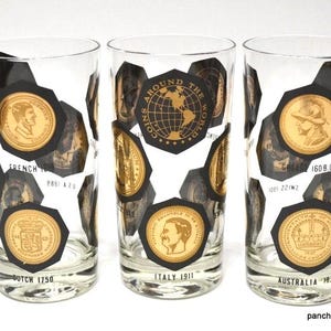 May include: Three clear glass tumblers decorated with black hexagonal shapes containing gold-colored coin illustrations. Each glass features text identifying the coins, such as French 1810, Dutch 1750, Italy 1911, Greece 160 B.C., and Australia 1937.