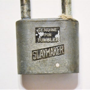 May include: Close-up of a vintage silver-colored padlock with the text "GENUINE PIN TUMBLER" and "SLAYMAKER" embossed on the front. The lock has a rectangular body and two metal loops at the top.
