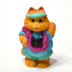 May include: A small orange cat figurine wearing a blue shirt, pink lei, and a blue visor. The cat is holding a black camera and waving with its right paw.