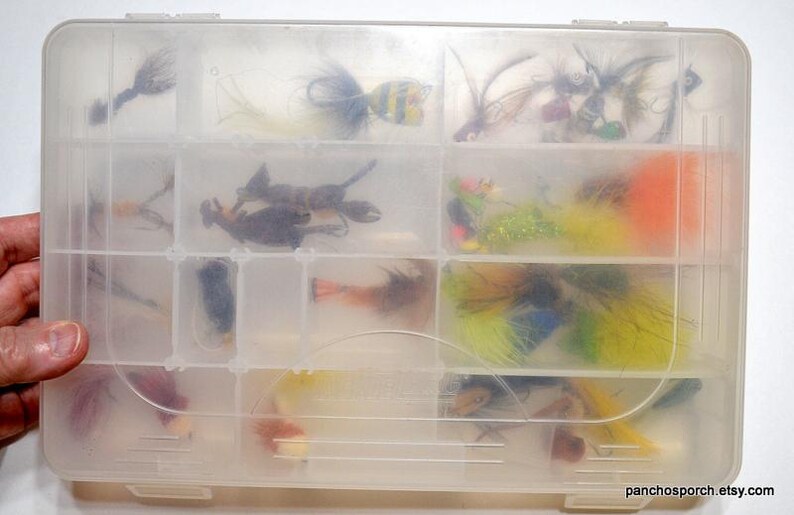 May include: A clear plastic tackle box filled with various fishing lures. The lures are colorful, with feathers, beads, and other materials. The box is divided into multiple compartments for organization.