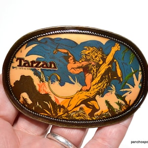May include: Vintage oval belt buckle featuring a Tarzan illustration. The buckle has a bronze-colored frame with a rope-like border. The image depicts Tarzan and a lion in a jungle setting, with the word "Tarzan" printed at the top.