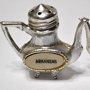 May include: A pair of vintage silver-colored salt and pepper shakers. The salt shaker is shaped like a teapot with the word "ARKANSAS" on the front. The pepper shaker is similar in design. Both have small feet and a handle.