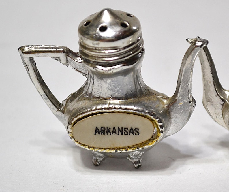 May include: A pair of vintage silver-colored salt and pepper shakers. The salt shaker is shaped like a teapot with the word "ARKANSAS" on the front. The pepper shaker is similar in design. Both have small feet and a handle.