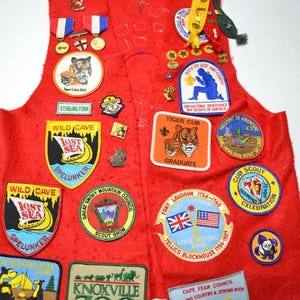 May include: A vintage red Boy Scout vest covered in patches and pins. Patches feature text like "Wild Cave," "Knoxville Zoo," and "Tiger Cub." Various pins and medals are also attached, showcasing a history of scouting.