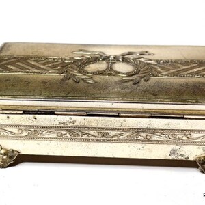 May include: An antique rectangular jewellery box with a hinged lid. The box is off-white with ornate silver-coloured metal accents. The lid features a decorative design with a central emblem and laurel leaves. The box rests on four decorative feet.