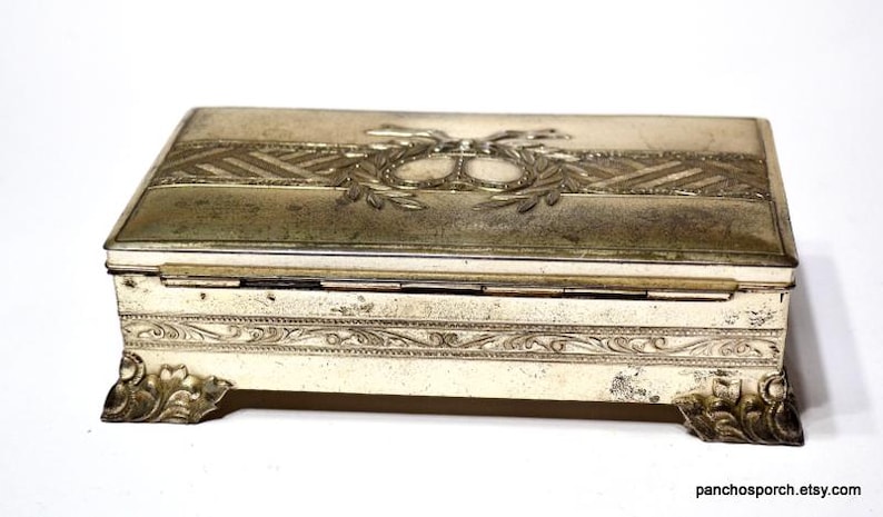 May include: An antique rectangular jewellery box with a hinged lid. The box is off-white with ornate silver-coloured metal accents. The lid features a decorative design with a central emblem and laurel leaves. The box rests on four decorative feet.