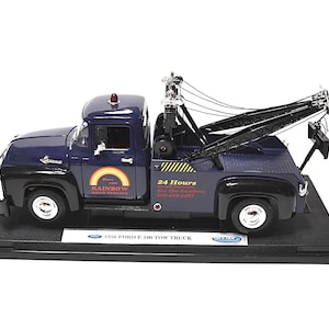 May include: A diecast model of a 1956 Ford F-100 tow truck. The truck is navy blue and black, with a tow crane and the text "Rainbow Road Service". The model sits on a black display base.