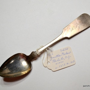 May include: Silver spoon with a rounded bowl and a rectangular handle. The handle is engraved with initials. A tag attached to the spoon reads "Jonathan Packard, Rochester, N.Y." and other handwritten details. The spoon is likely vintage.
