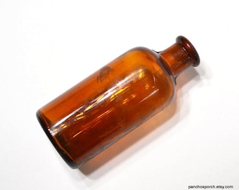 May include: A small, cylindrical amber glass bottle with a smooth, rounded body and a narrow, short neck featuring a thick, flared rim. The translucent brown glass has a glossy finish and reflects light across its curved surface.