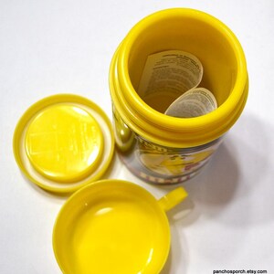 May include: A bright yellow container with a matching lid and small cup. The container is open, revealing a folded paper inside. The cup has a small handle. The container has a printed design around the middle.