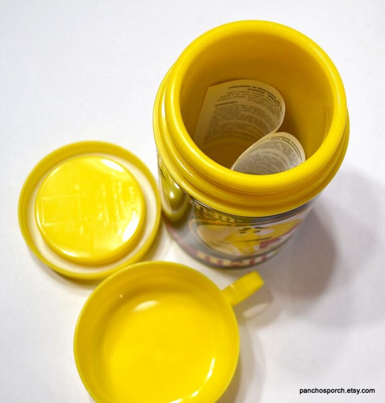 May include: A bright yellow container with a matching lid and small cup. The container is open, revealing a folded paper inside. The cup has a small handle. The container has a printed design around the middle.