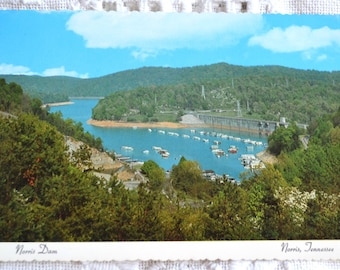 May include: Vintage postcard featuring a scenic view of Norris Dam in Tennessee. The image showcases a large body of water surrounded by lush green trees and a dam structure. The sky is a clear blue, and the postcard has a scalloped edge.