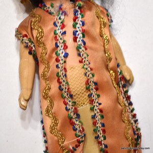 Puede incluir: Una mu&ntilde;eca vintage con un vestido color melocot&oacute;n con adornos dorados y detalles coloridos. El vestido est&aacute; adornado con detalles rojos, azules y verdes. Se ven los brazos y las piernas de la mu&ntilde;eca, y tiene pelo negro.
