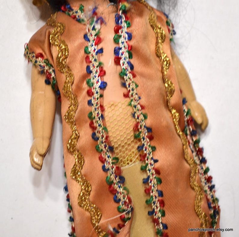Puede incluir: Una mu&ntilde;eca vintage con un vestido color melocot&oacute;n con adornos dorados y detalles coloridos. El vestido est&aacute; adornado con detalles rojos, azules y verdes. Se ven los brazos y las piernas de la mu&ntilde;eca, y tiene pelo negro.
