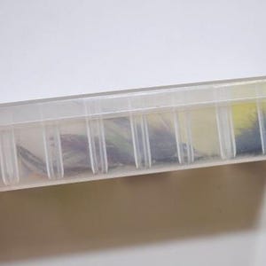 May include: A clear plastic fishing tackle box with a hinged lid and multiple compartments. The box contains various fishing lures in shades of orange, yellow, blue, and black. The box is held by a hand.