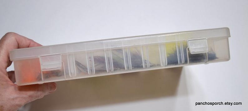 May include: A clear plastic fishing tackle box with a hinged lid and multiple compartments. The box contains various fishing lures in shades of orange, yellow, blue, and black. The box is held by a hand.