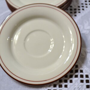 May include: A stack of off-white saucers with a thin, reddish-brown border. The saucers have a recessed center for a cup and are arranged on a white tablecloth.