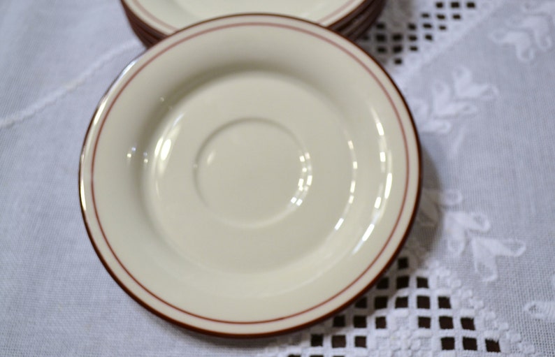 May include: A stack of off-white saucers with a thin, reddish-brown border. The saucers have a recessed center for a cup and are arranged on a white tablecloth.