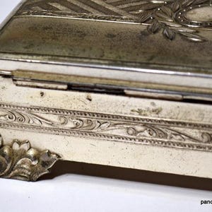 May include: An antique silver-toned rectangular box with ornate detailing. The box has a hinged lid with engraved patterns and a decorative border. The corners feature stylized floral accents. The box has a vintage, aged appearance.
