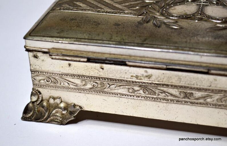 May include: An antique silver-toned rectangular box with ornate detailing. The box has a hinged lid with engraved patterns and a decorative border. The corners feature stylized floral accents. The box has a vintage, aged appearance.