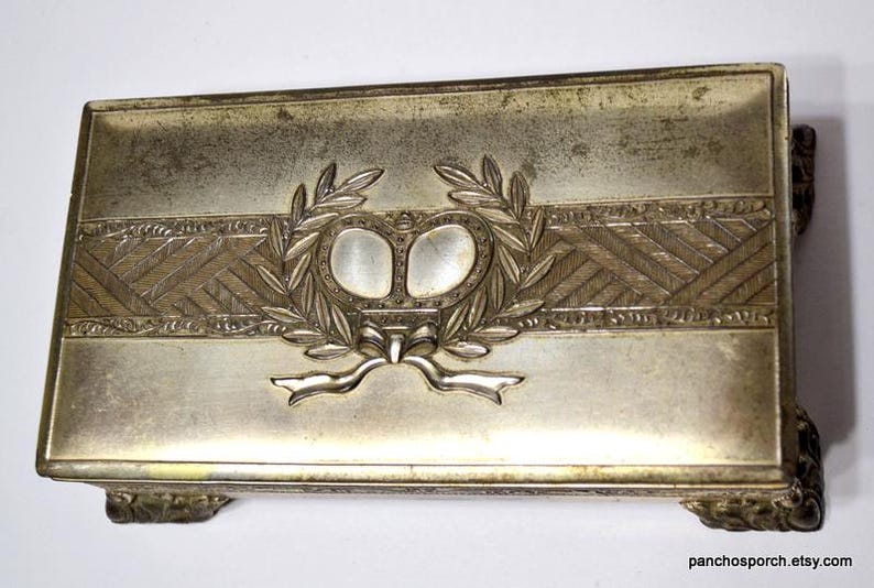 May include: Silver-toned rectangular box with decorative features. The box has a central band with a laurel wreath and a bow design. The corners have decorative feet. The box has an aged, antique appearance.