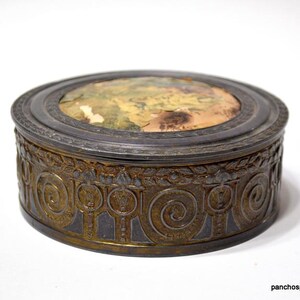 Op de afbeelding: Een antieke, ovale sieradendoos met een gedetailleerde, bronskleurige buitenkant. Het deksel heeft een vervaagde, kleurrijke afbeelding. De doos heeft een donkere, metalen rand en een decoratief, herhalend patroon rond de basis.