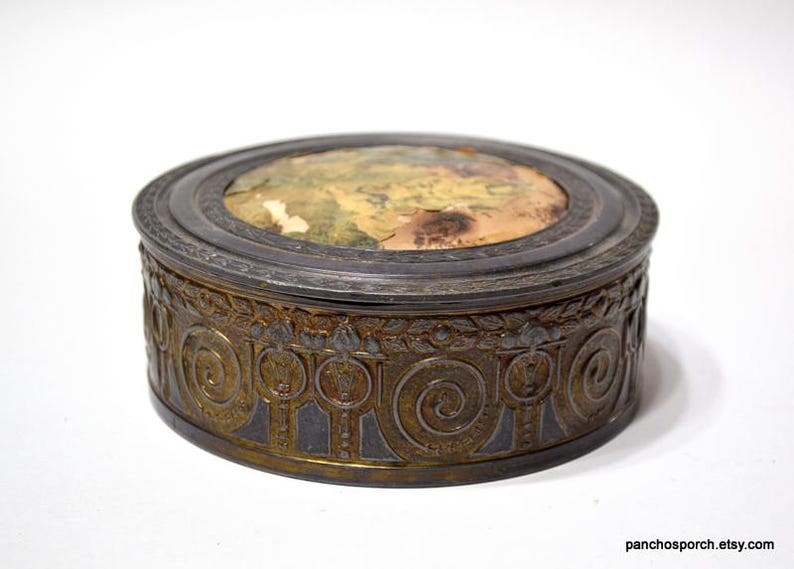 Op de afbeelding: Een antieke, ovale sieradendoos met een gedetailleerde, bronskleurige buitenkant. Het deksel heeft een vervaagde, kleurrijke afbeelding. De doos heeft een donkere, metalen rand en een decoratief, herhalend patroon rond de basis.