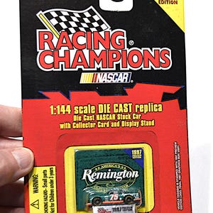 May include: A 1997 Racing Champions die-cast NASCAR replica of a Remington car. The packaging is red and black, with the Racing Champions and NASCAR logos. The car is green and white, with the number 75. Includes a collector card and display stand.