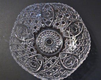 May include: A clear glass bowl featuring an intricate pressed pattern with a central sunburst design surrounded by alternating diamond-shaped motifs and cross-hatched textures. The circular bowl has a scalloped rim and a reflective surface.