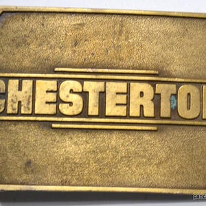 May include: A vintage brass belt buckle with the name "CHESTERTON" in raised letters. The buckle has a rectangular shape with rounded edges and a tarnished, aged appearance. The text is centered on the buckle.