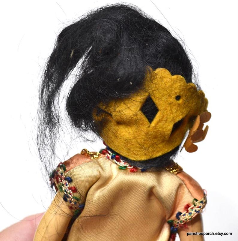 Puede incluir: Una mu&ntilde;eca vintage con cabello negro y una corona dorada. La mu&ntilde;eca lleva un vestido color beige con detalles bordados coloridos en las mangas. El cabello de la mu&ntilde;eca est&aacute; peinado lejos de la cara.