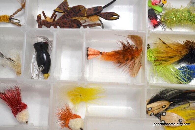 May include: A clear plastic tackle box filled with various fishing lures. The lures include a black lure, brown crab-shaped lures, and colorful feathered lures in shades of red, orange, yellow, and green.