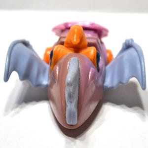 May include: A colorful toy dragon with a pink body, orange accents, and light blue wings. The dragon has a light blue crest and a pink headpiece. The toy is set against a white background.