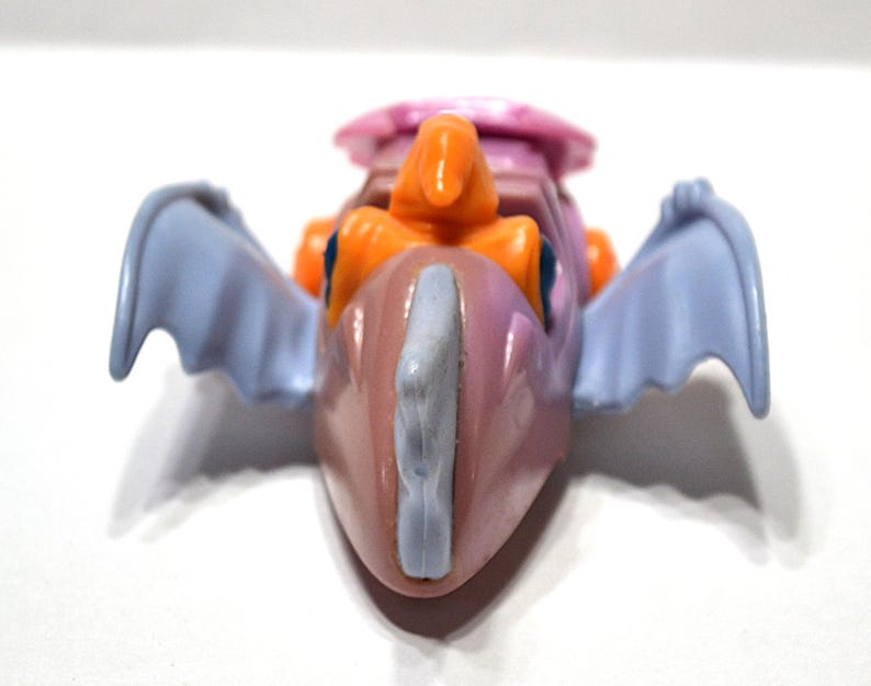 May include: A colorful toy dragon with a pink body, orange accents, and light blue wings. The dragon has a light blue crest and a pink headpiece. The toy is set against a white background.