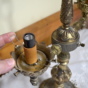 Puede incluir: Una l&aacute;mpara de metal en tono lat&oacute;n con una columna central ornamentada con motivos de hojas y varios casquillos. Cada casquillo est&aacute; rodeado por una base festoneada en forma de p&eacute;talo e incluye un clip de alambre para sujetar la pantalla.
