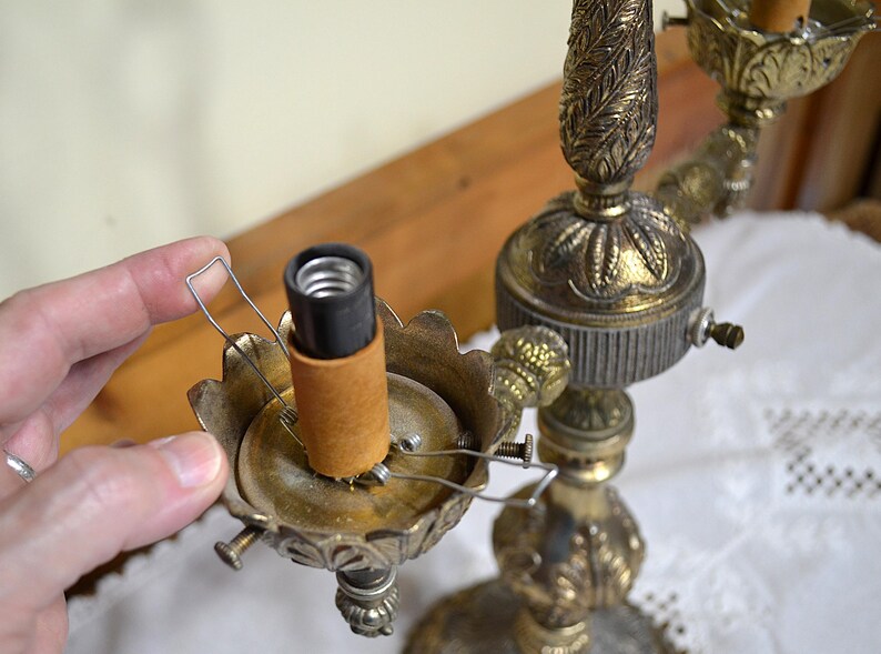 Puede incluir: Una l&aacute;mpara de metal en tono lat&oacute;n con una columna central ornamentada con motivos de hojas y varios casquillos. Cada casquillo est&aacute; rodeado por una base festoneada en forma de p&eacute;talo e incluye un clip de alambre para sujetar la pantalla.