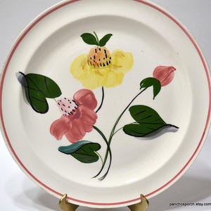 May include: A white ceramic plate with a pink rim and floral design. The hand-painted flowers are yellow and pink with green leaves and black stems. The plate is a vintage piece.