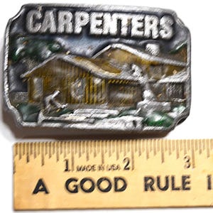 May include: A silver-colored belt buckle with the word "CARPENTERS" at the top. The buckle features a detailed relief of a house and a person. A wooden ruler with the text "A GOOD RULE" is below the buckle.