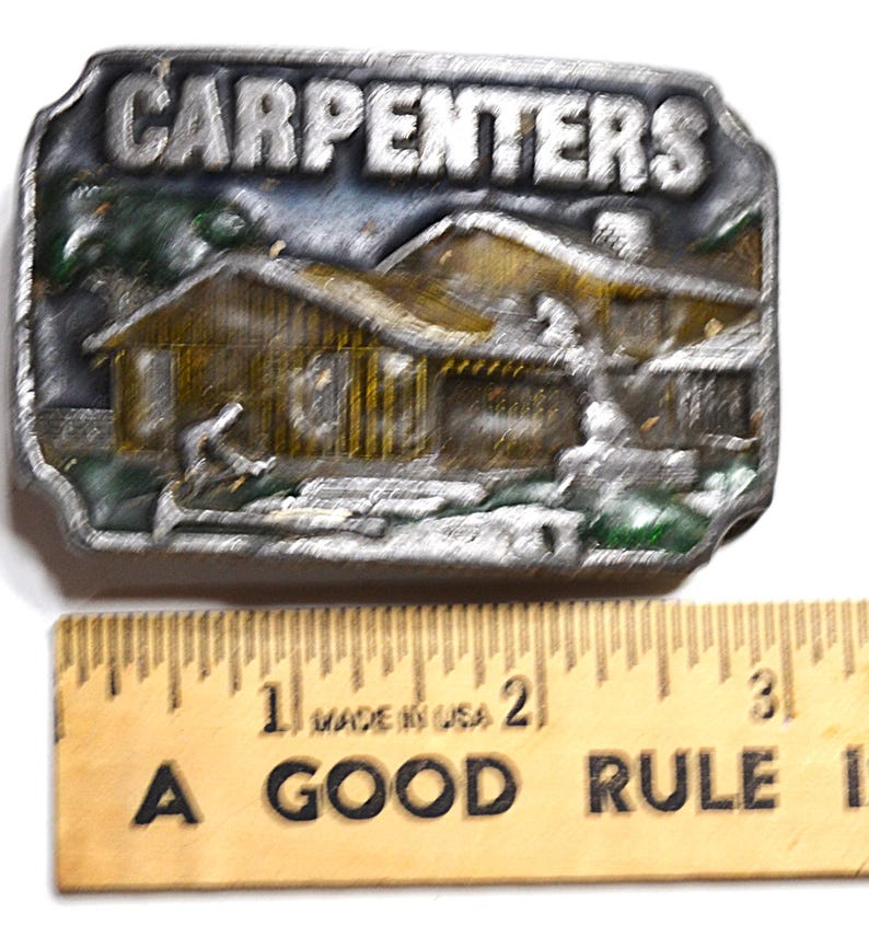 May include: A silver-colored belt buckle with the word "CARPENTERS" at the top. The buckle features a detailed relief of a house and a person. A wooden ruler with the text "A GOOD RULE" is below the buckle.